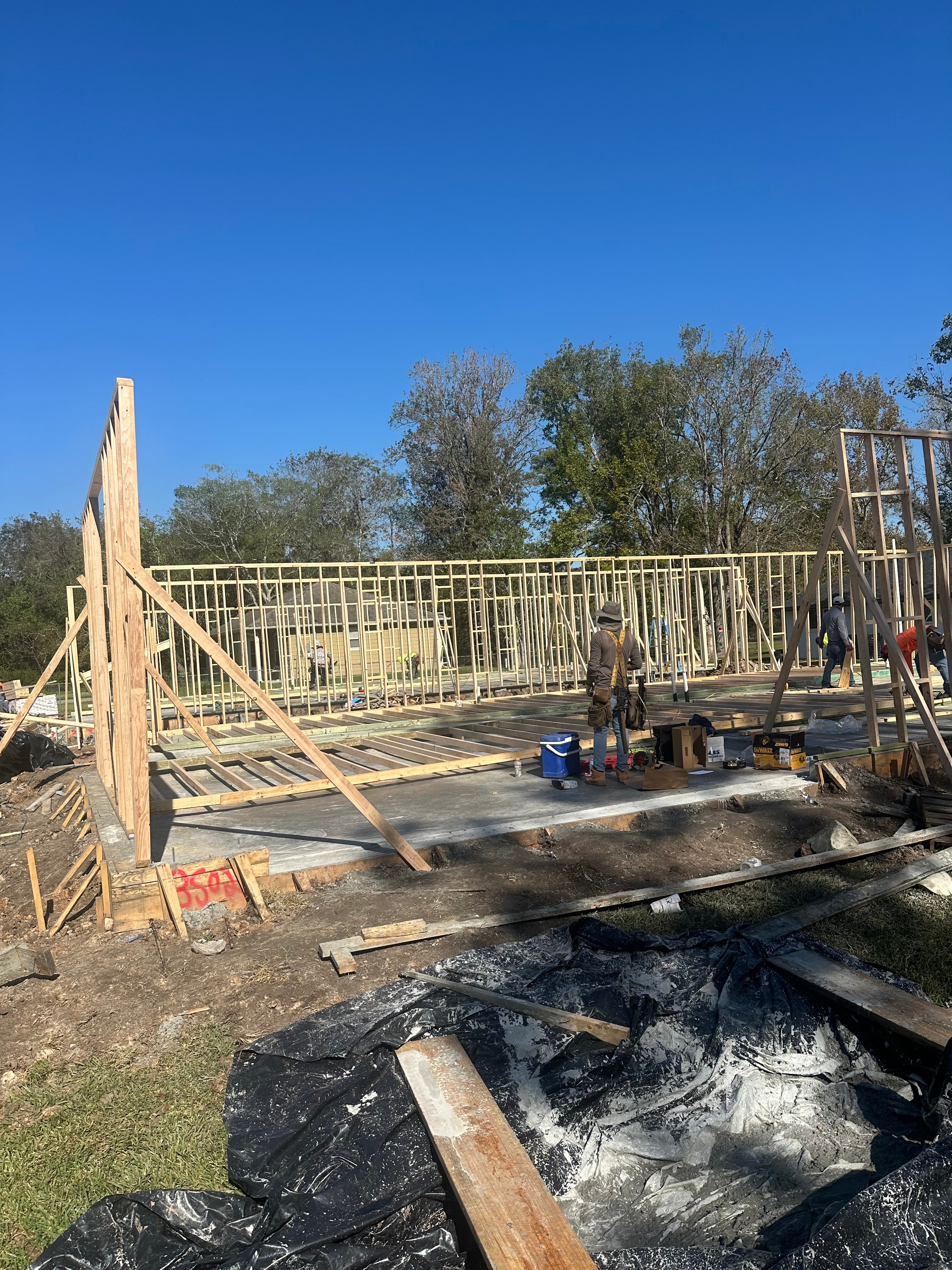 Worker installs wooden wall framing on a concrete foundation under a clear blue sky.